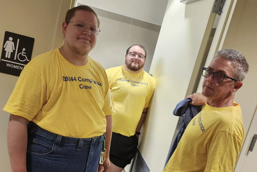 Three trans masculine activists with the Compliance Crew stand at the door of a public women's restroom.