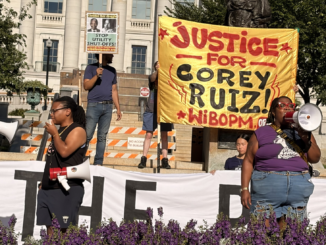 Two protesters in front of "Justice for Corey Ruiz" banner speak into megaphones.